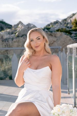 A figure in a white dress poses against a backdrop of mountains and a glass railing in an outdoor setting.