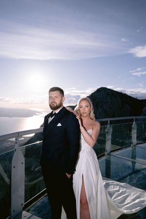 A couple in wedding attire poses on a scenic mountain-top observation deck at sunset.