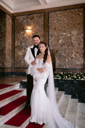 A wedding couple poses on red carpeted stairs in an elegant venue with stone walls and wall sconce lighting.