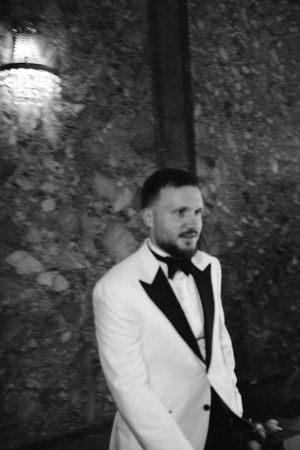A person in a white dinner jacket with black lapels and bow tie poses against a textured stone wall at night.