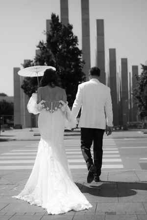 A couple in wedding attire walks along a modern plaza with tall pillars, the bride holding a white parasol.
