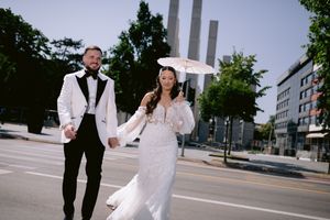 A wedding couple walks hand in hand on a city street with modern buildings in the background.