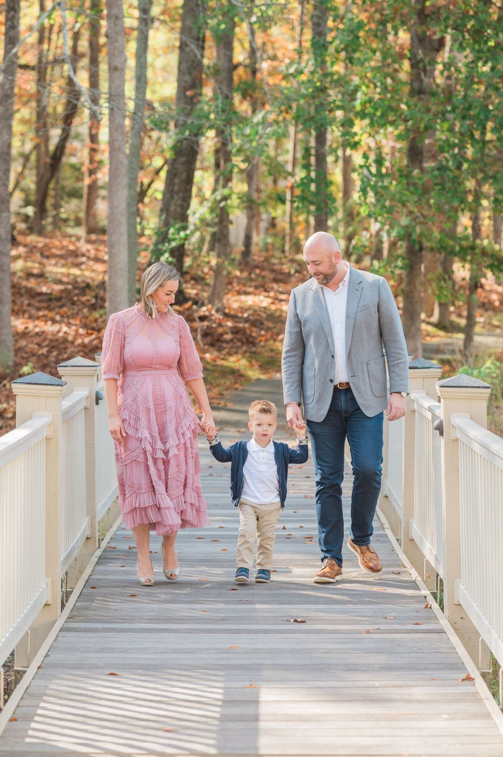 A family walks together across a white wooden bridge surrounded by autumn trees in the forest.