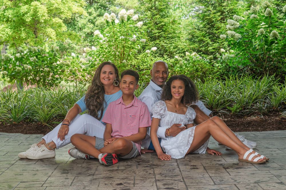 Family group poses together outdoors in a garden setting wearing coordinated white and pink casual summer outfits.