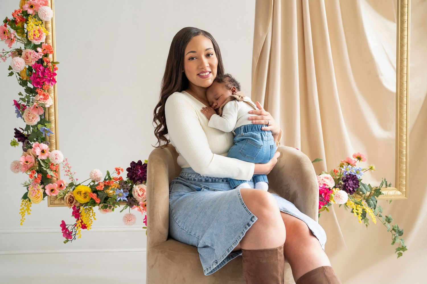 Mother and baby share tender moment in elegant studio setting with floral decorations and cream curtains.