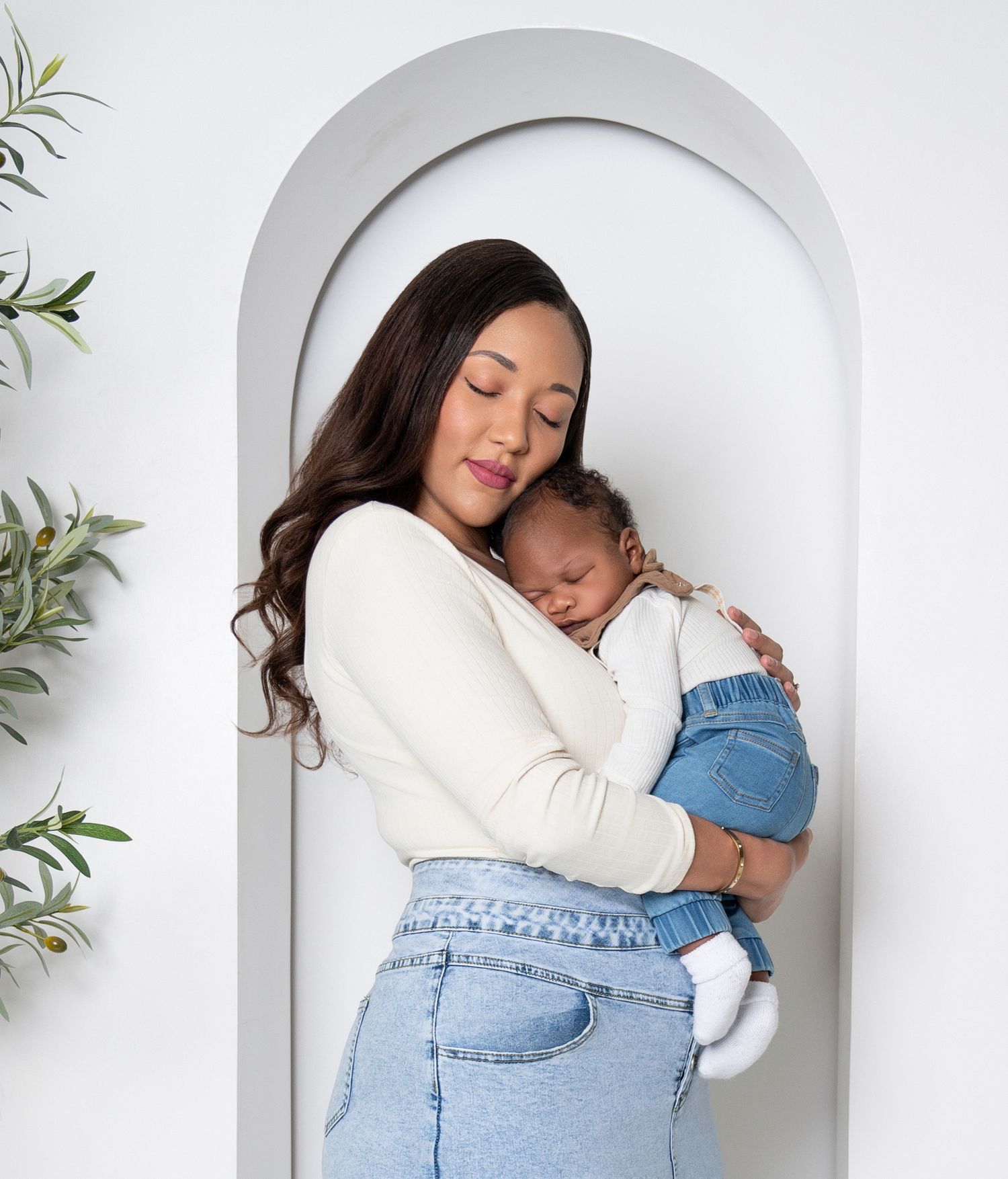 Parent embraces sleeping infant in white sweater against minimalist archway with plants.