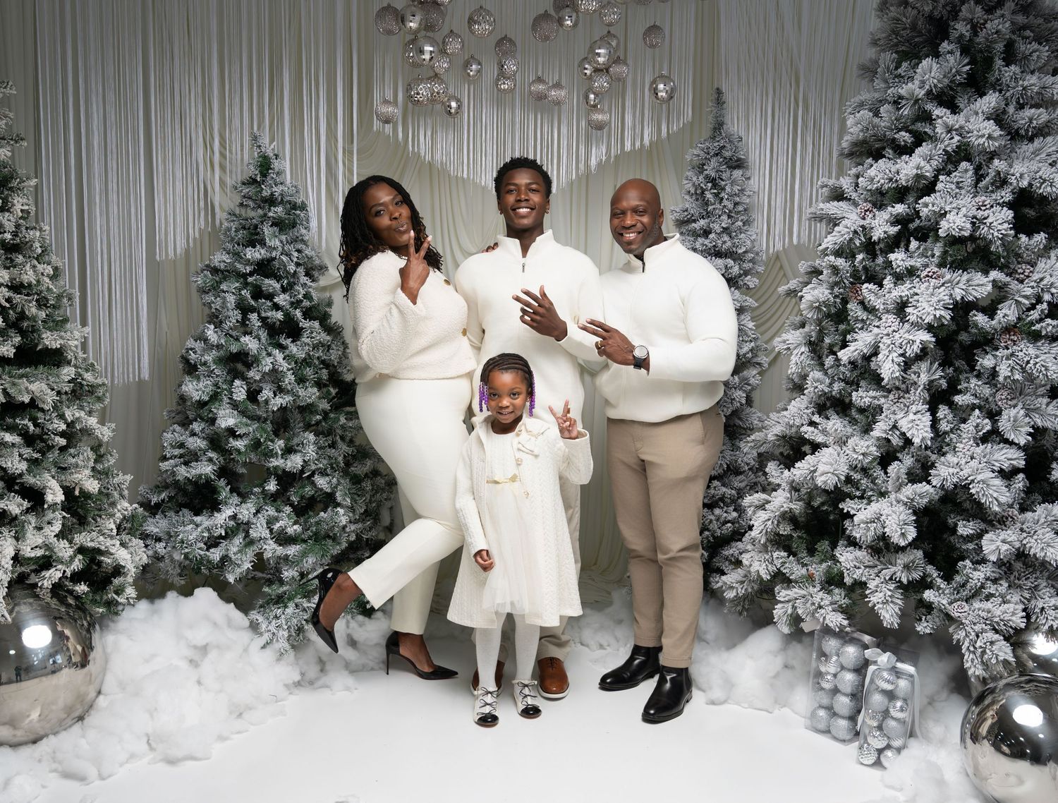 Family poses for holiday photos in front of snowy Christmas trees wearing coordinated white and tan winter outfits.
