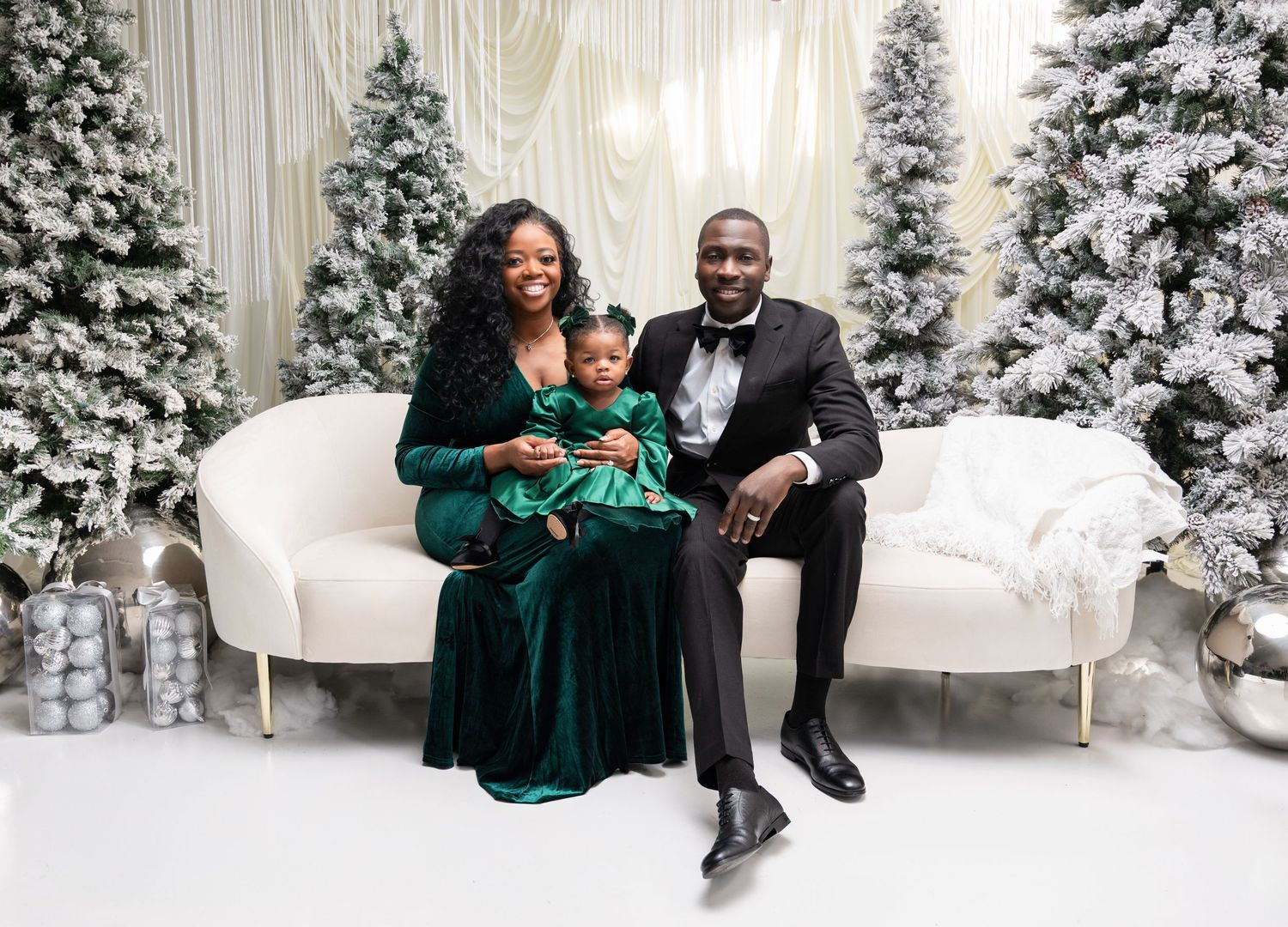 A family wearing green velvet and black outfits poses together in a winter wonderland setting with snow-covered trees.