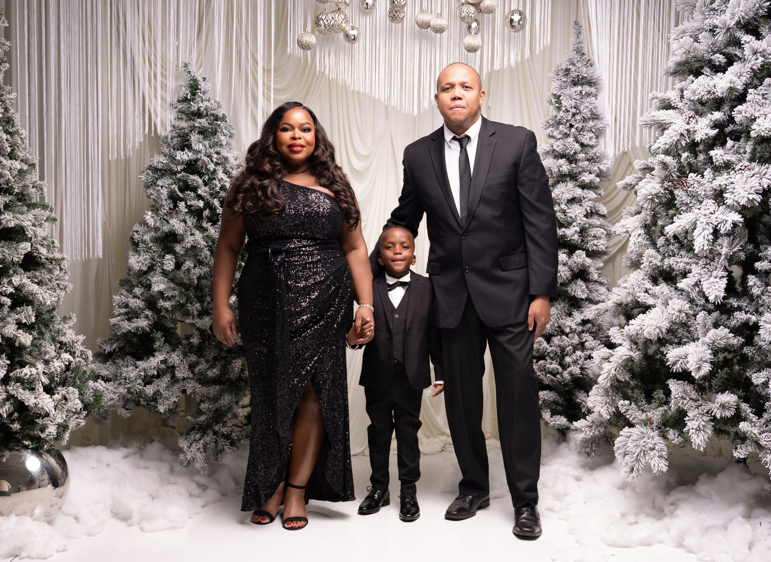 Elegant family poses in formal attire against a winter wonderland backdrop with snow-covered Christmas trees and white curtains.