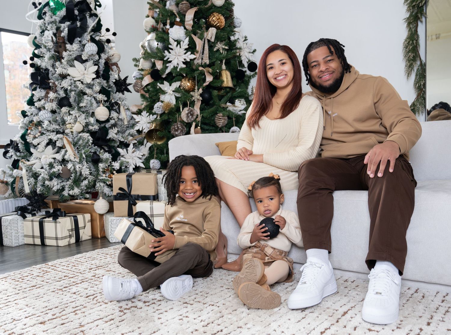 Family portrait in matching neutral colors sitting together on a white couch near Christmas decorations.