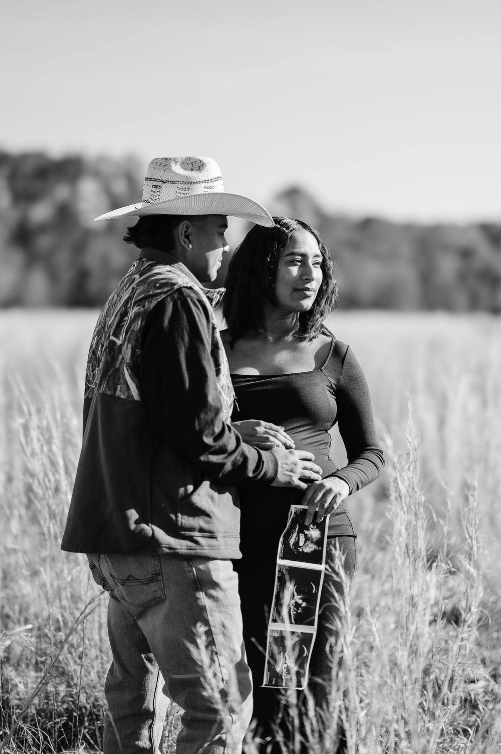 A romantic black and white portrait photograph of a couple standing together in a sunlit field of tall grass.
