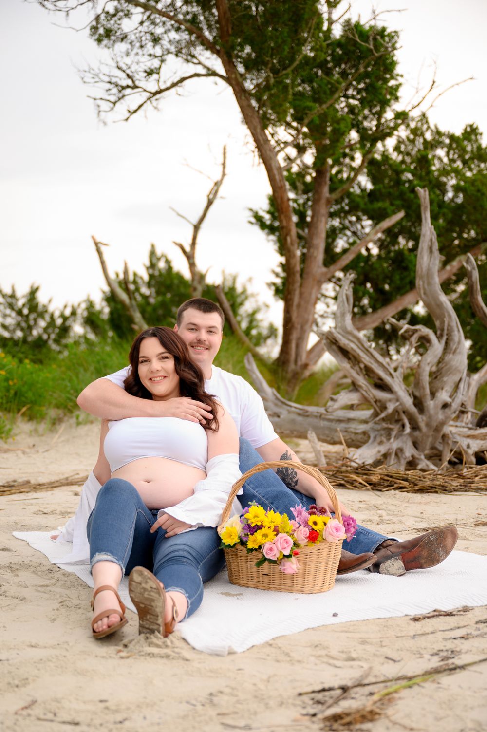 Expecting couple relaxes on beach blanket with flower basket near driftwood.