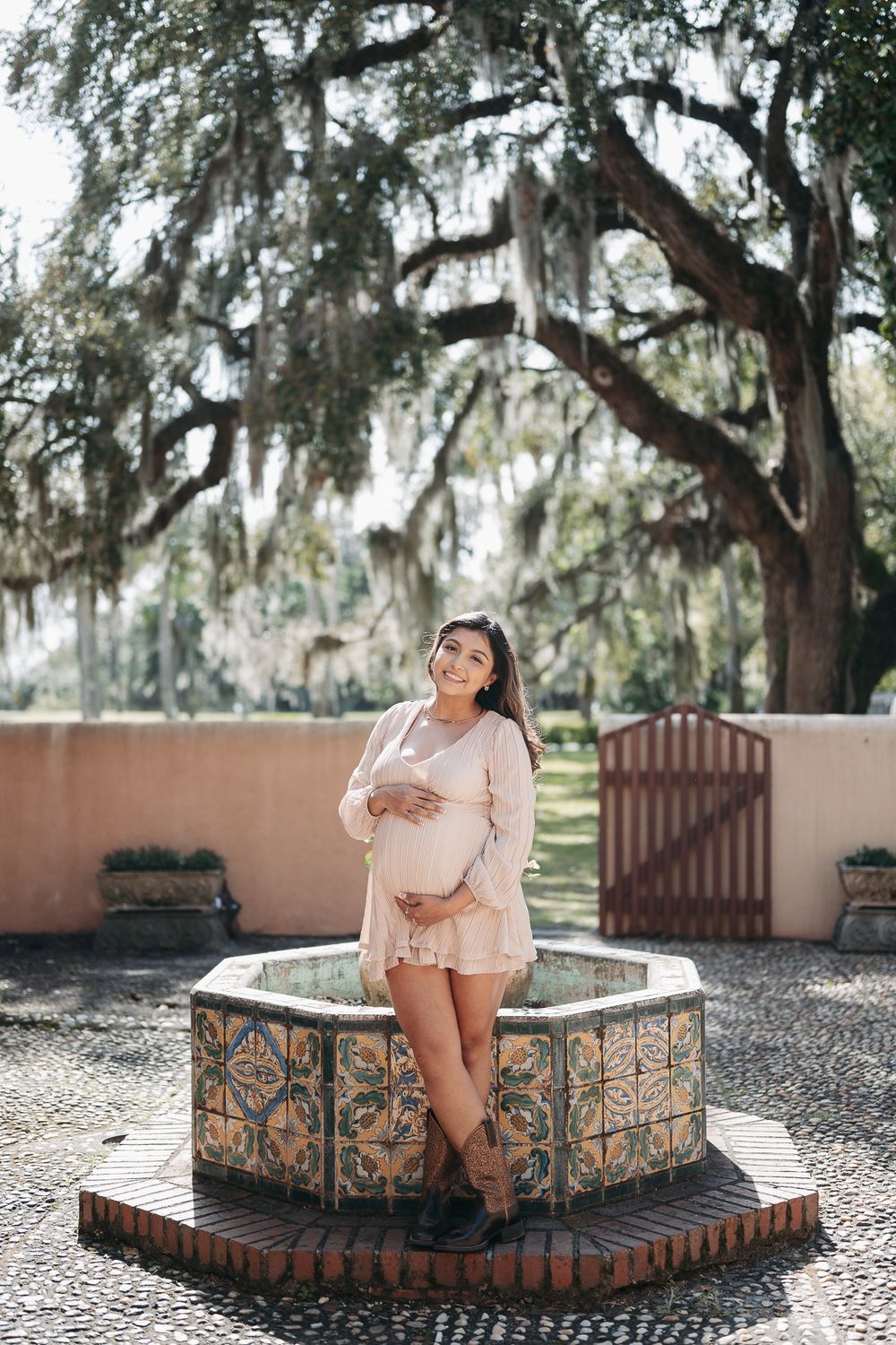 A pregnant woman stands by a decorative stone fountain under Spanish moss draped oak trees in the South.
