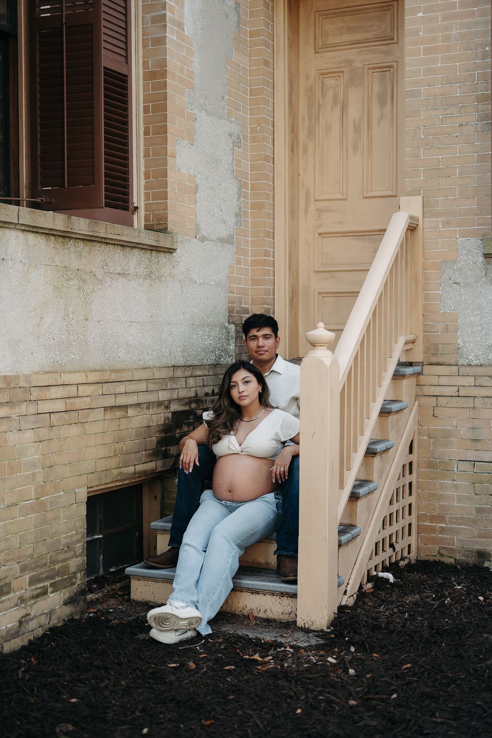 Couple sitting on outdoor staircase against brick wall during maternity photoshoot.