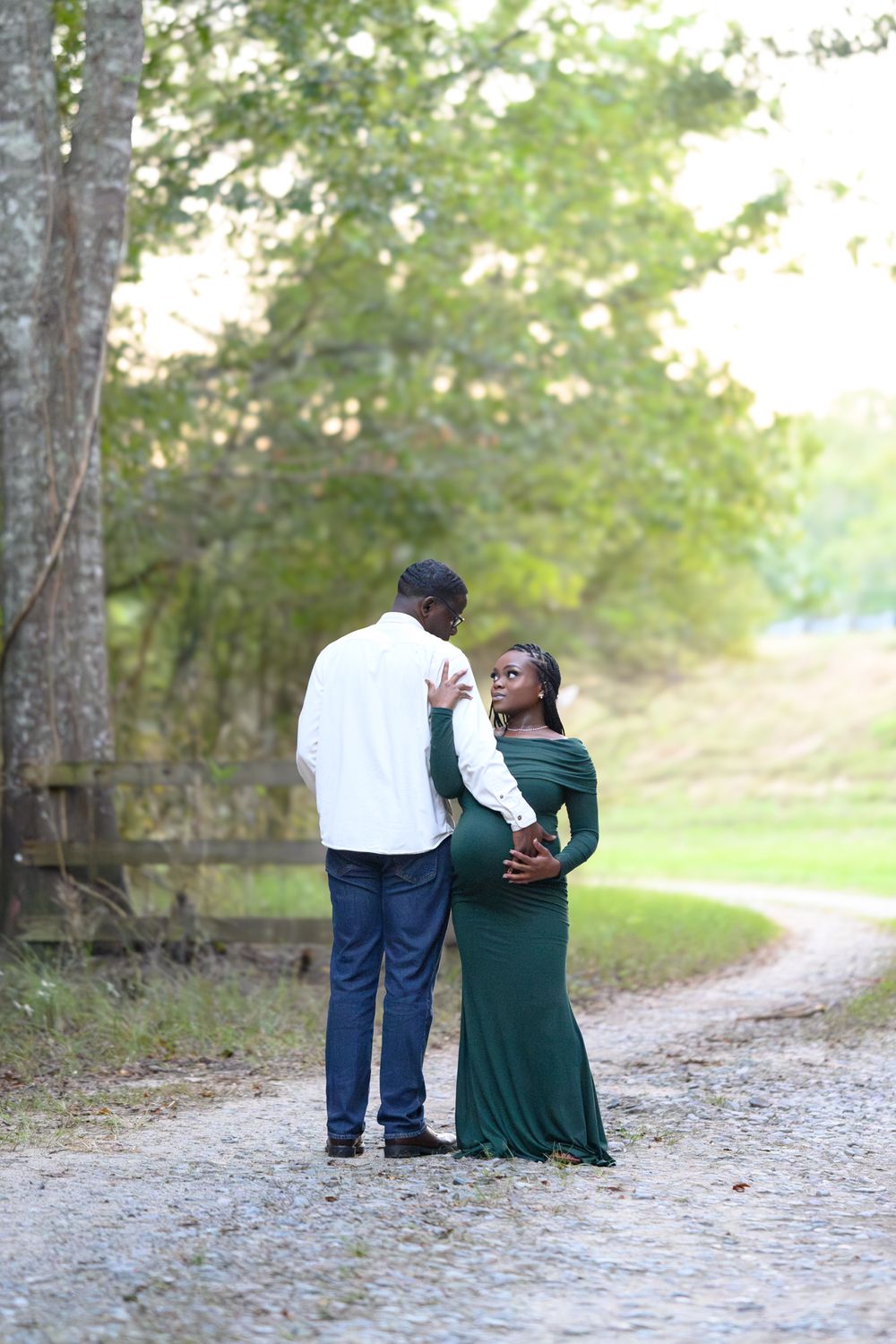 A couple walks along a tree-lined path, sharing a tender moment during their maternity photoshoot.