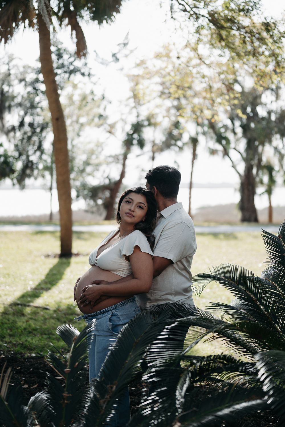 A couple embraces in a sunlit garden with palm fronds, capturing a romantic maternity moment among the trees.