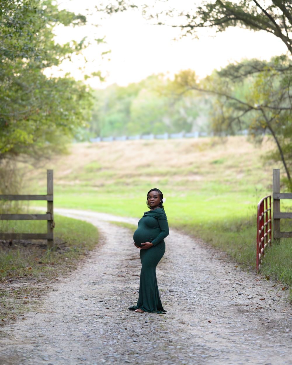 Expecting mother in a green dress poses gracefully on a winding path framed by wooden fences and trees.