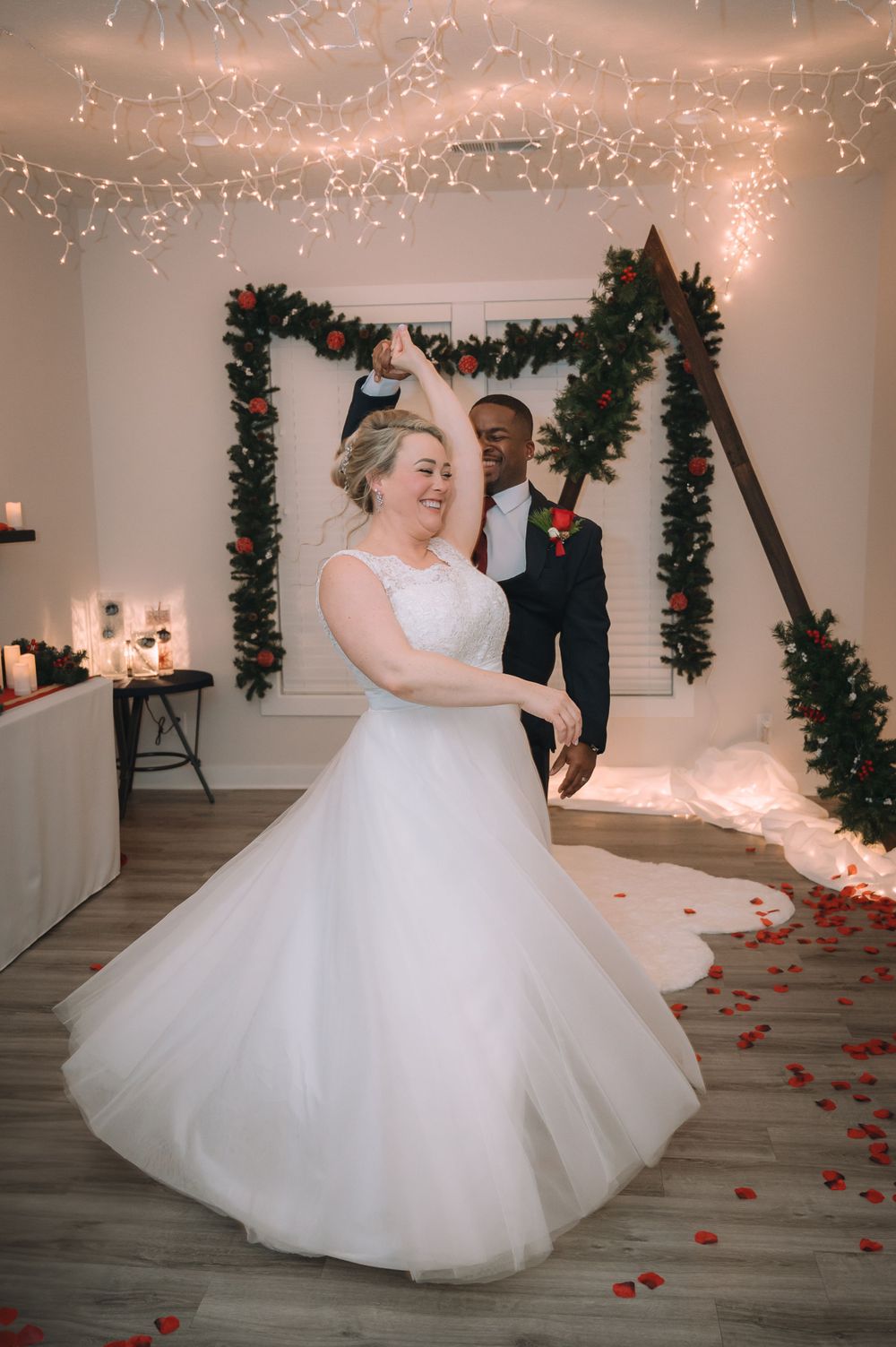 A couple shares a joyful dance moment under twinkling lights and greenery garland.