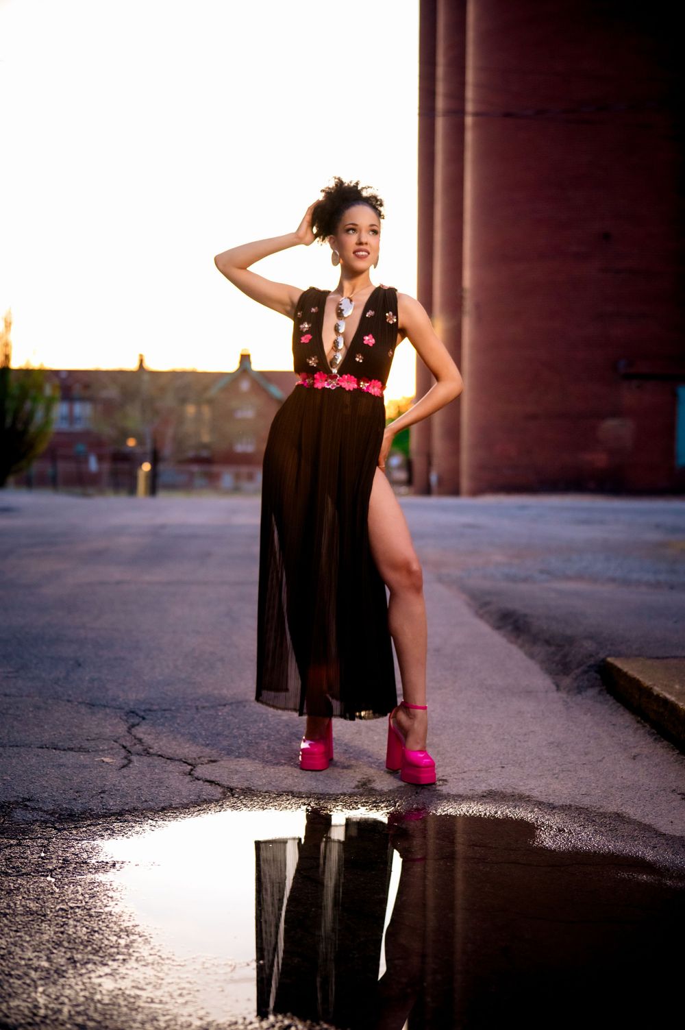 A dancer in a black dress and pink high heels performs elegantly near a puddle on an urban street at sunset.