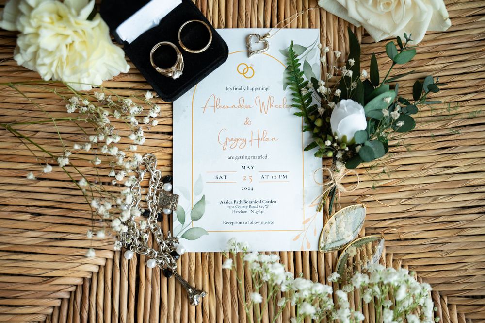 Wedding invitation with gold details surrounded by white flowers and eucalyptus on a woven surface.
