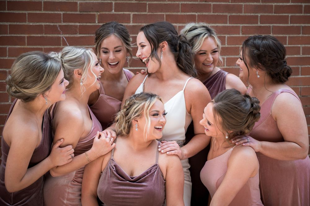 Bridesmaids in mauve dresses share a joyful moment against a brick wall.