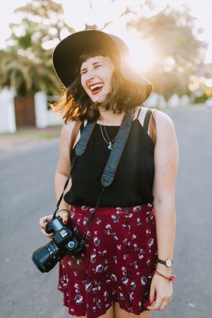 Photographer in sun backlit shot wearing black hat, dark tank top and floral burgundy shorts holding camera on street.