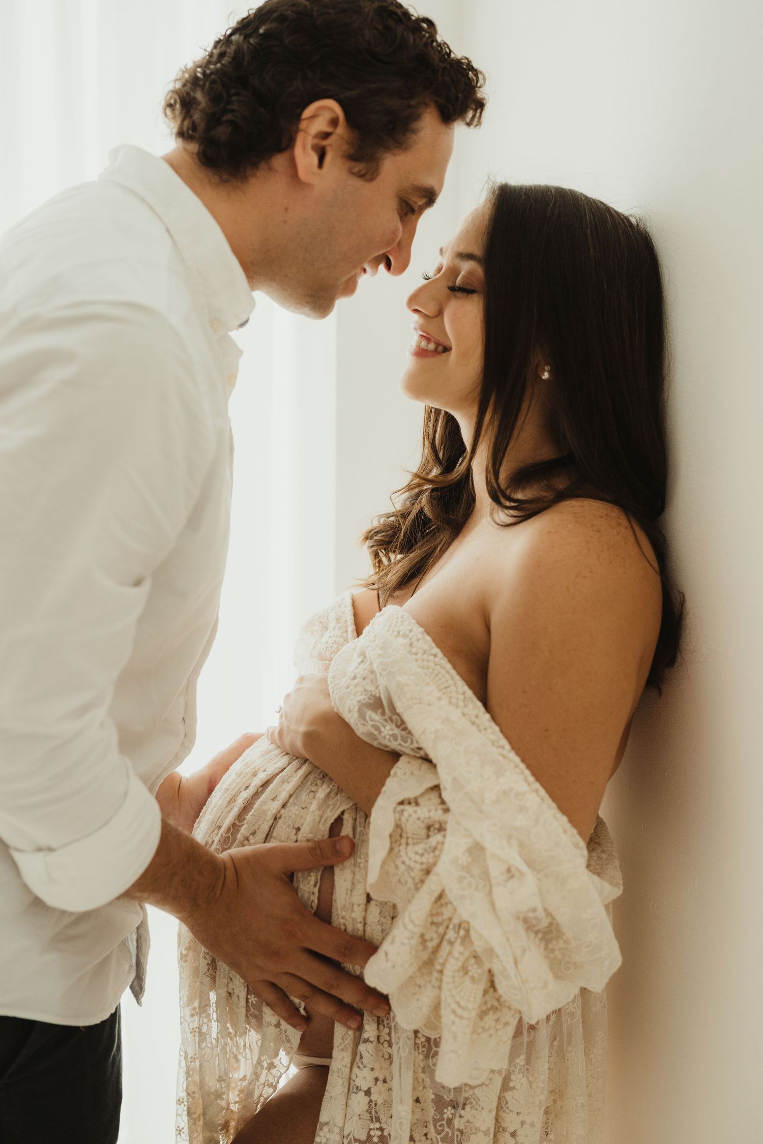 Intimate couple embracing near window in soft natural light during maternity photoshoot.