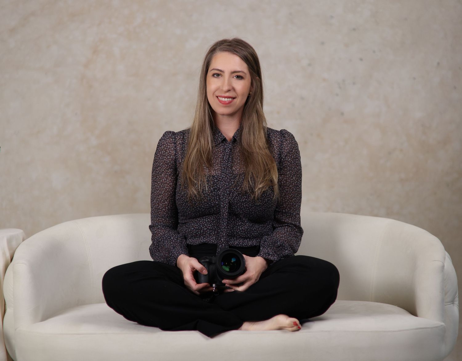 Someone in a dark sweater sits cross-legged on a white couch holding a camera, against a neutral background.