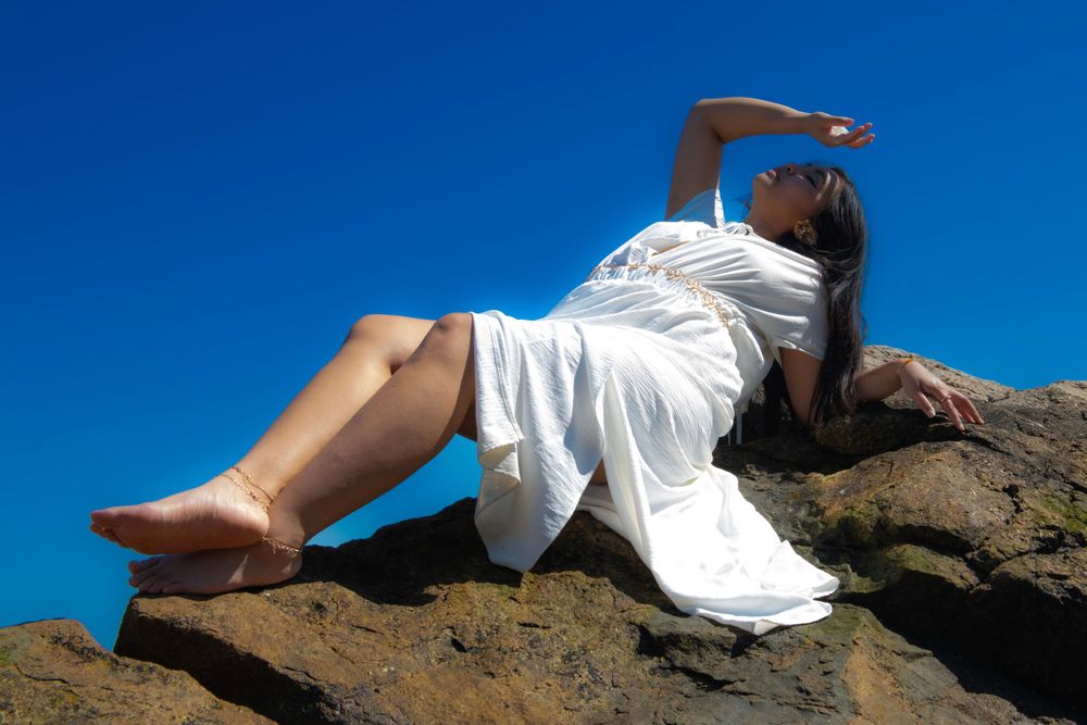 A person in white reclines artistically on rocky coastal terrain under blue sky.