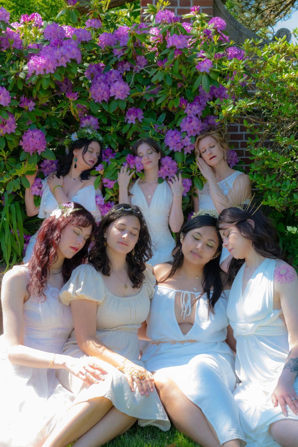 A group poses dreamily in white dresses against blooming purple rhododendrons.