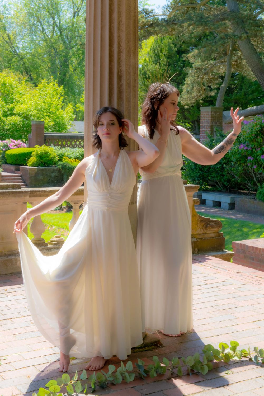 Two women in flowing white dresses pose gracefully against classical columns in a garden setting.