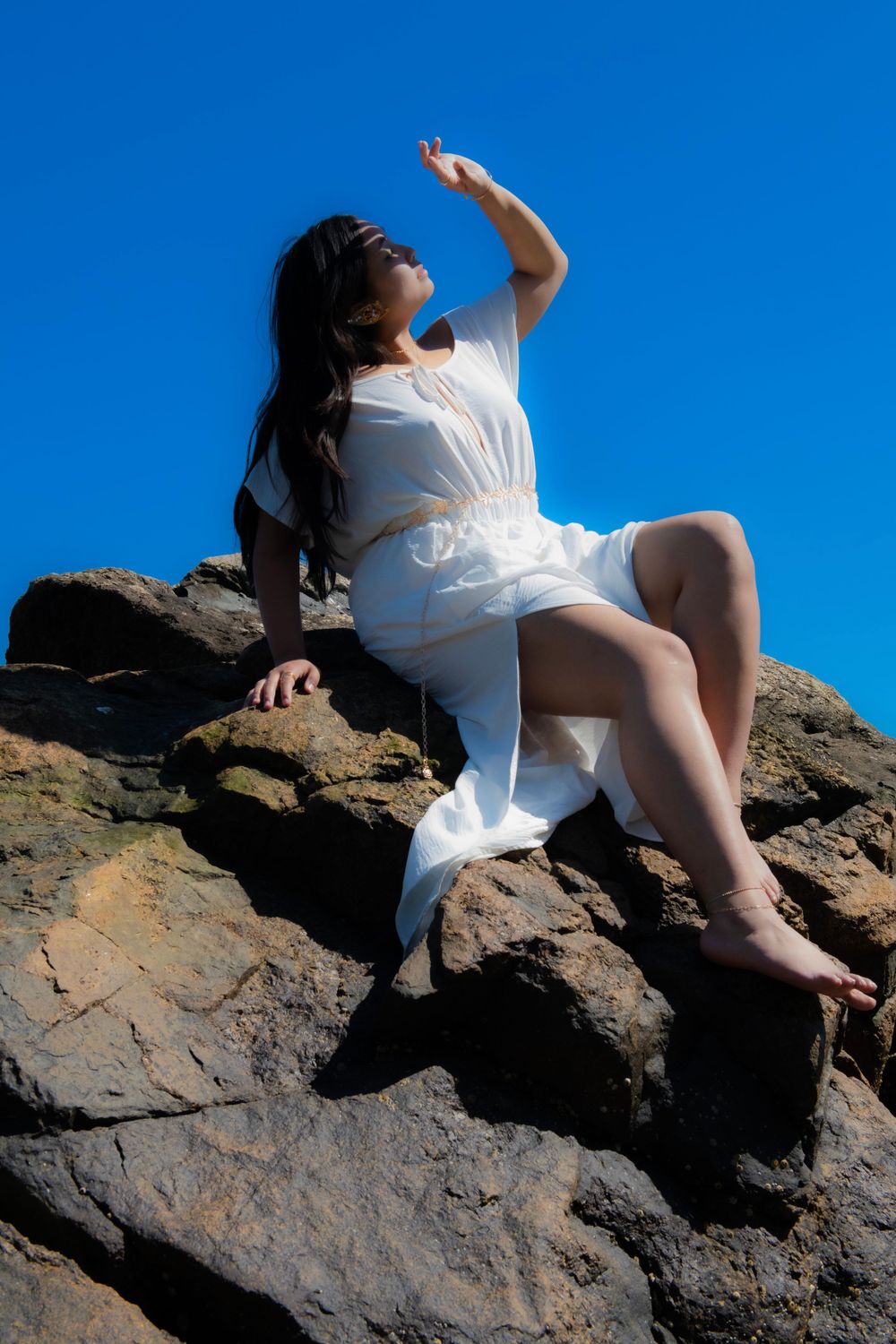 A figure in a white dress poses dramatically on coastal rocks against a bright blue sky.