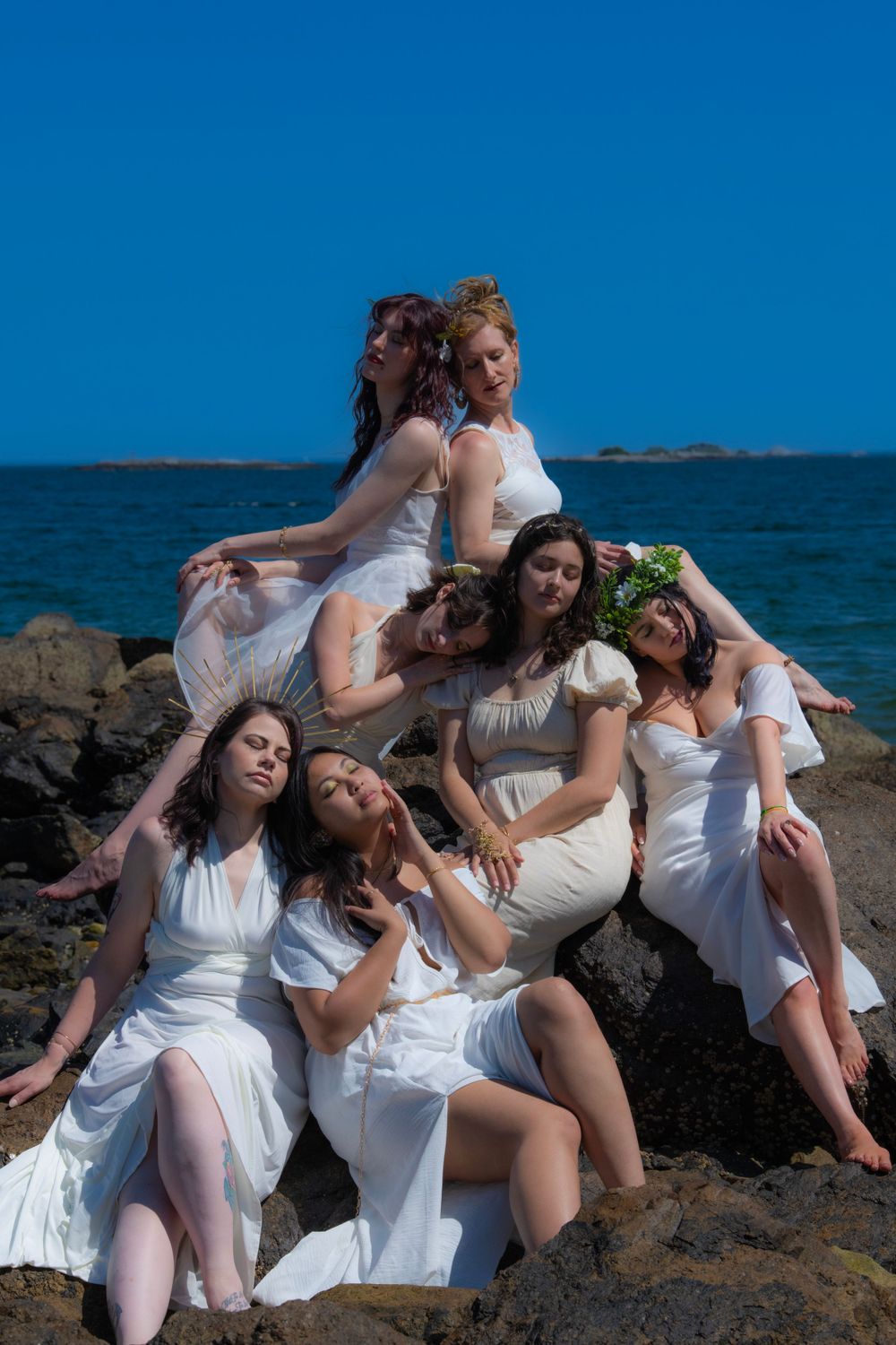 A group in white dresses poses artistically on oceanside rocks during a sunny photoshoot.