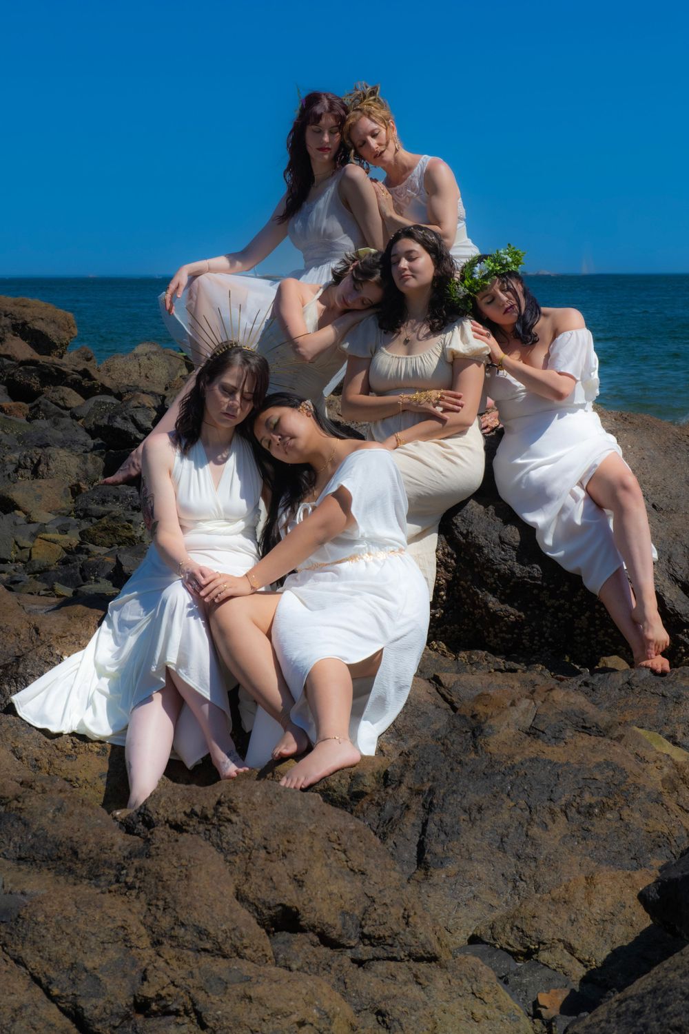 Friends in white dresses create an artistic group pose on coastal rocks by the sea.