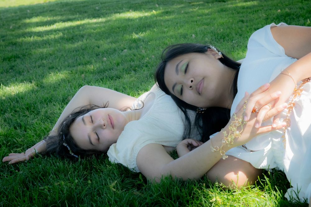 Two people in white dresses lay peacefully on green grass in dappled sunlight.