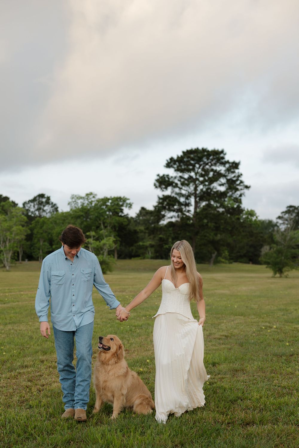 Couple holding hands in a green field with their golden retriever, woman in white dress, man in blue shirt.