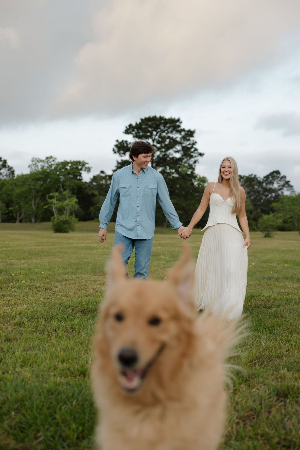 Couple holding hands walking in green field with golden retriever dog in foreground on cloudy day.