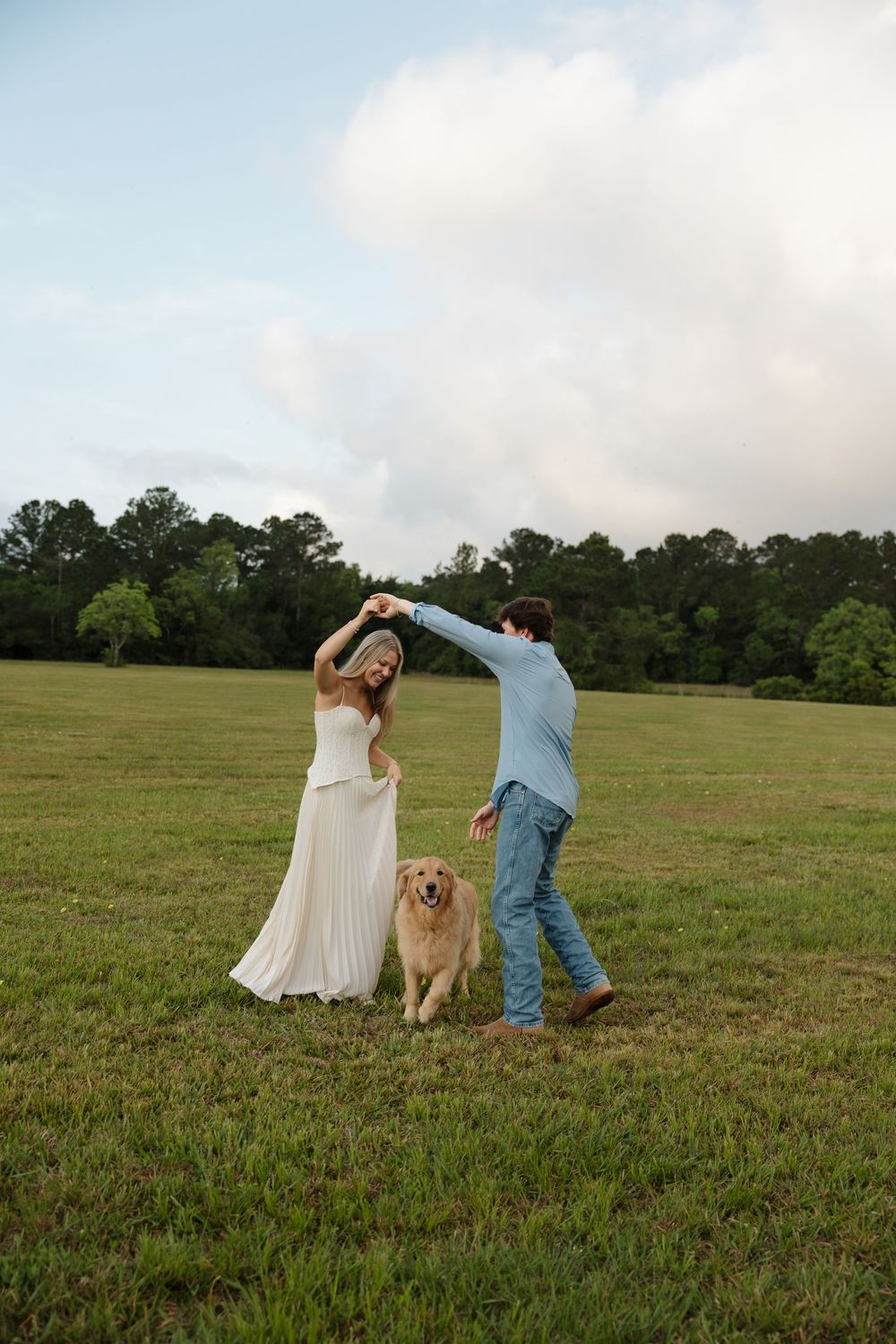 Couple dancing in a green field with a golden retriever, woman in white dress, man in blue shirt and jeans.