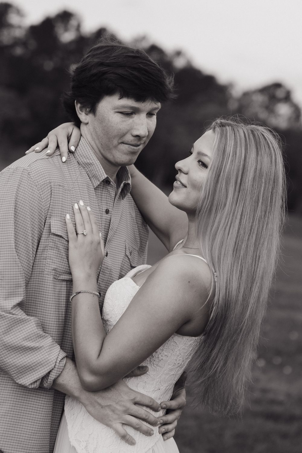 Black and white photo of a couple embracing outdoors, woman smiling with engagement ring visible, trees in background.