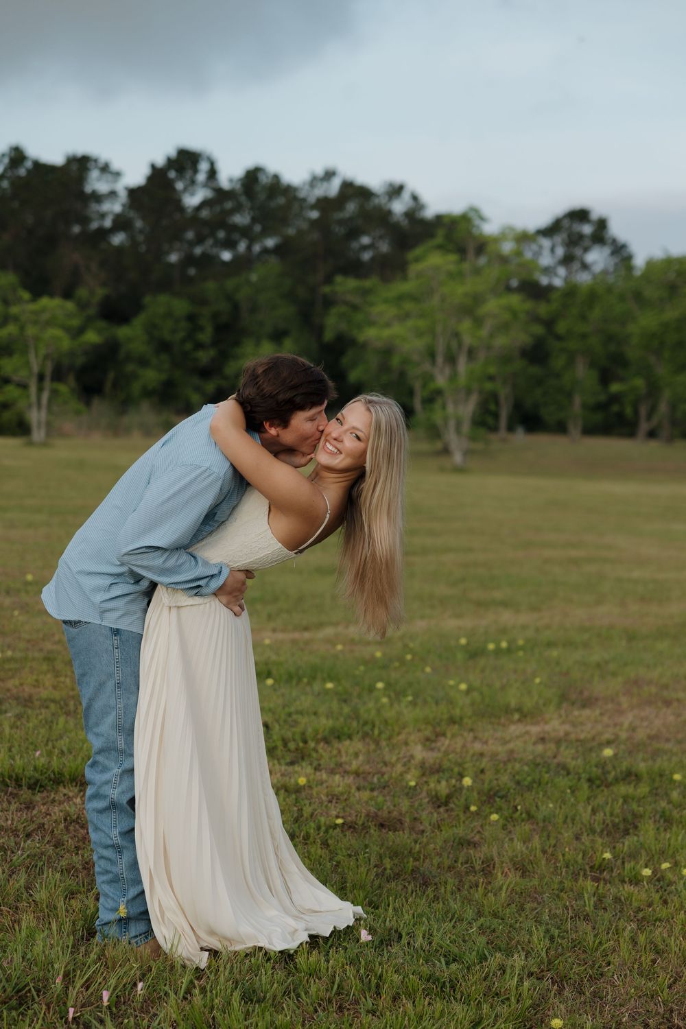 Couple sharing a romantic embrace in a green meadow, man kissing woman in white maxi dress outdoors.