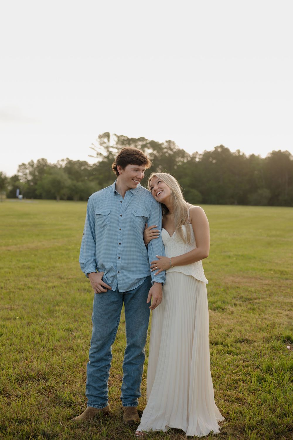 Happy couple embracing in a sunlit field, woman in white dress leaning on man in blue denim shirt and jeans.