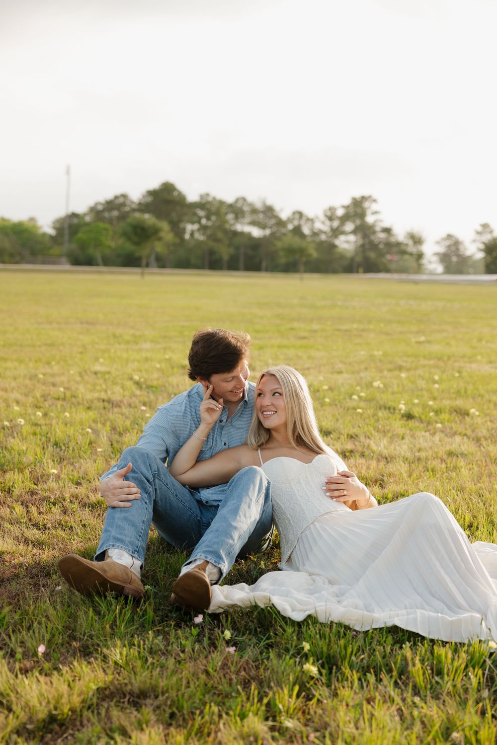 Couple sitting together on grass field, man whispering to smiling blonde woman in white dress.