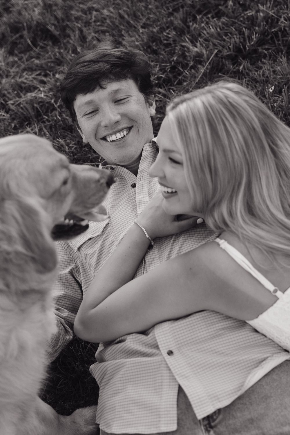 Black and white photo of laughing couple lying in grass with golden retriever licking the woman.