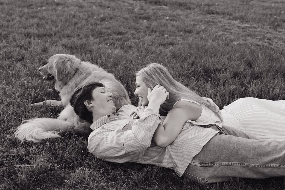 Couple lying on grass gazing at each other with a golden retriever dog resting beside them in black and white photo.