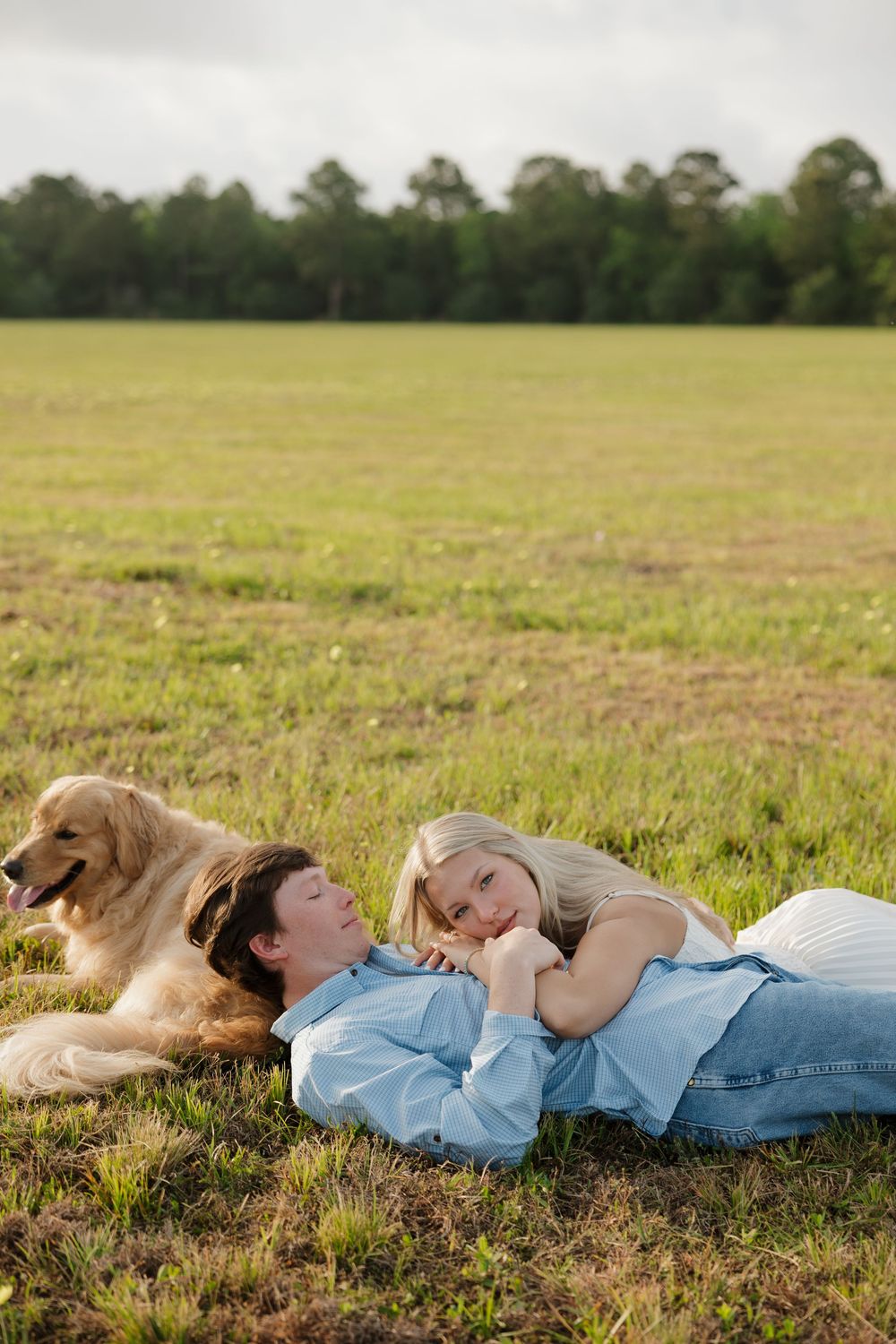 A couple relaxes on green grass in a field with their golden retriever dog on a cloudy day.