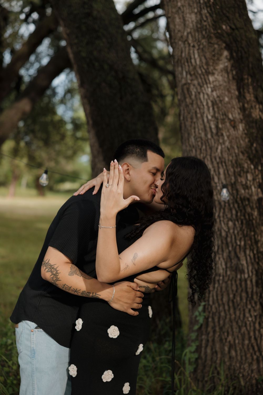 Couple sharing a romantic kiss near a tree in a park, woman wearing a floral black dress, man with tattoos.