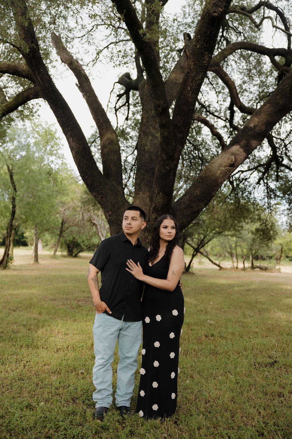 Couple posing together in front of a large oak tree in a green park, woman in black floral dress, man in black shirt.