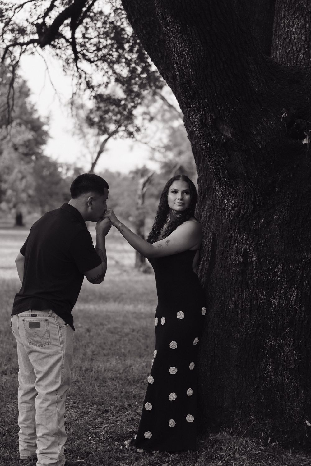 Black and white photo of a man kissing a woman's hand near a large tree in a park.