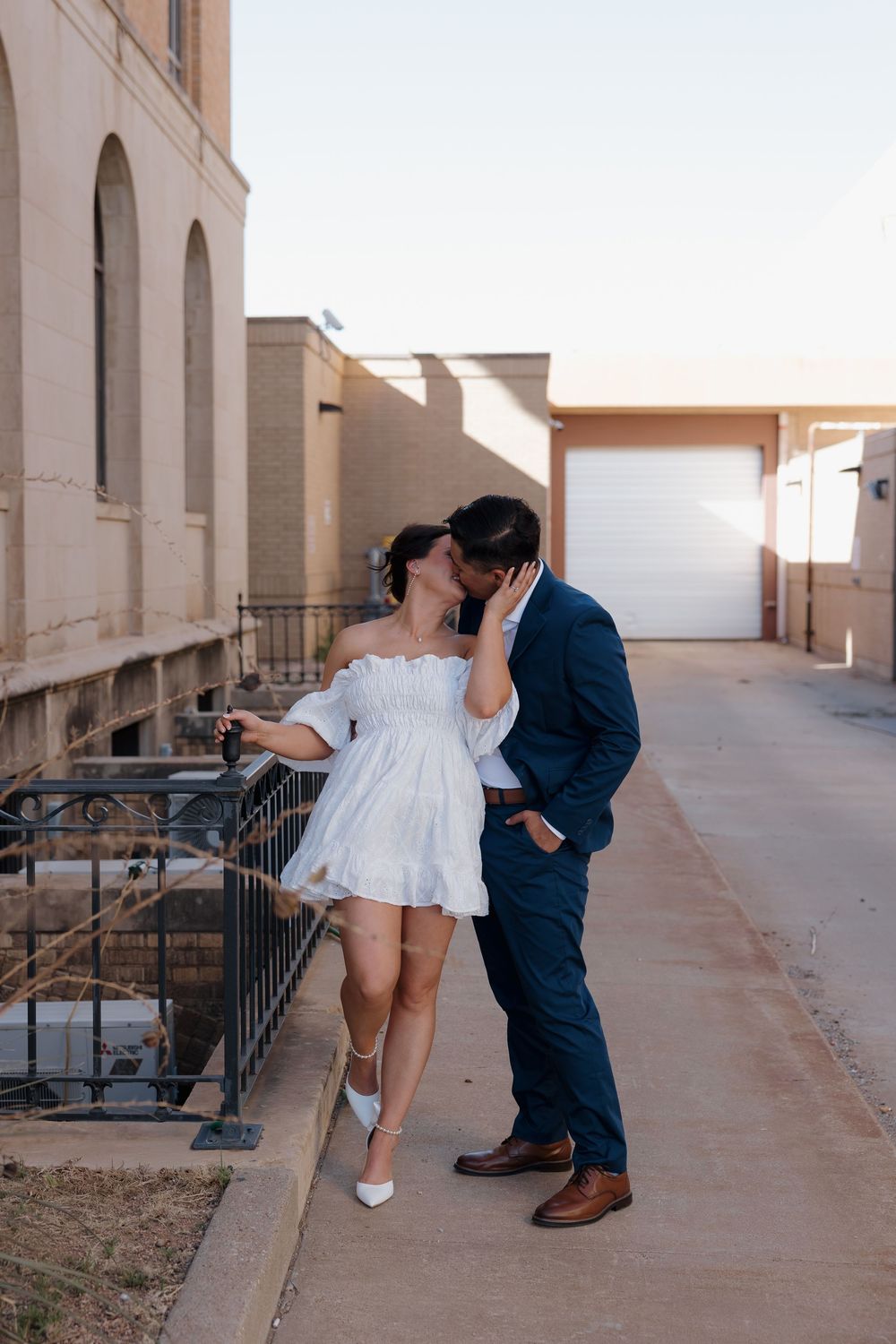 Couple shares a kiss on urban balcony at sunset, woman in white mini dress, man in navy suit.