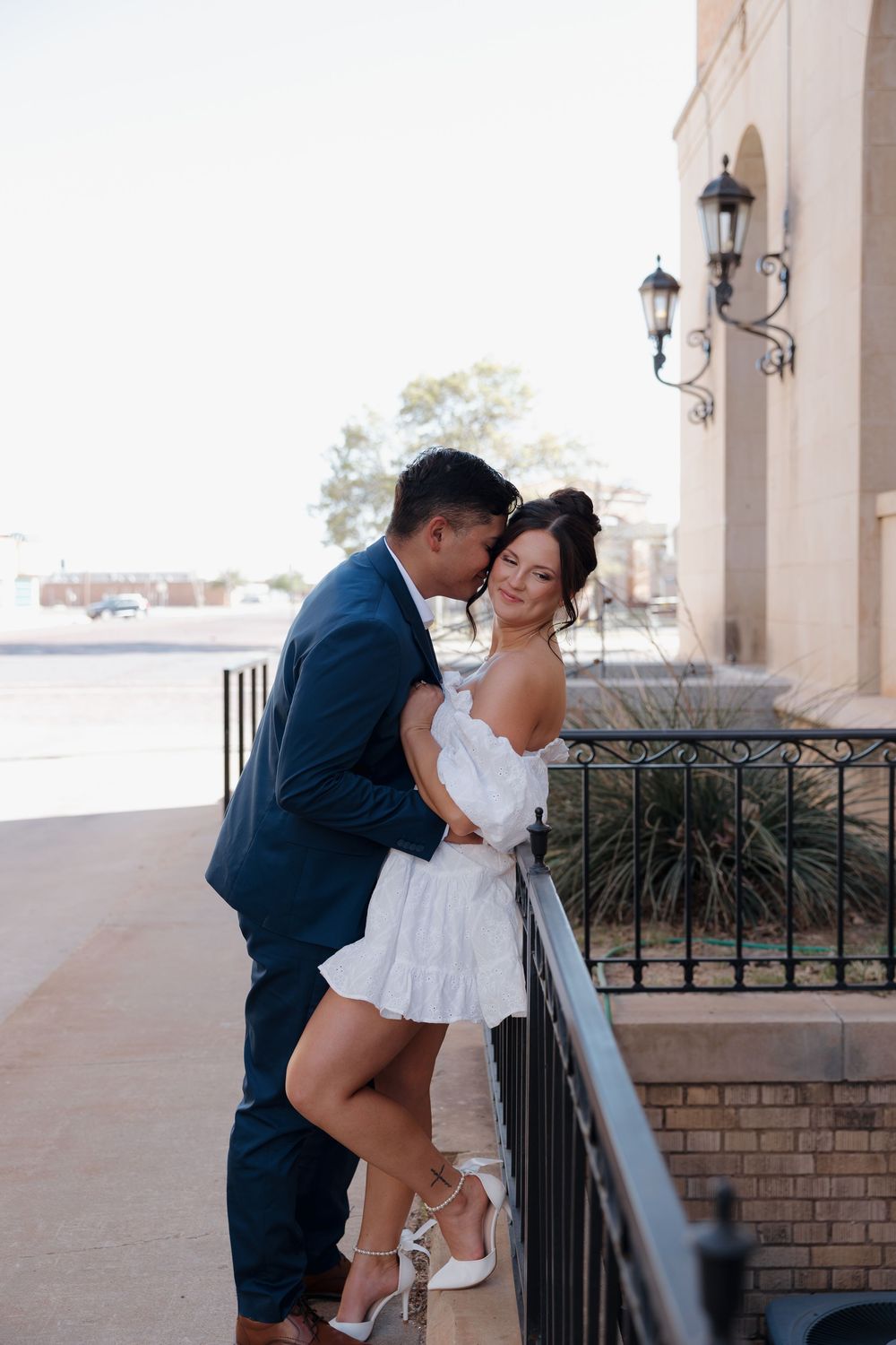 Couple embraces on elegant iron railing balcony, woman in white off-shoulder dress smiling warmly at camera.